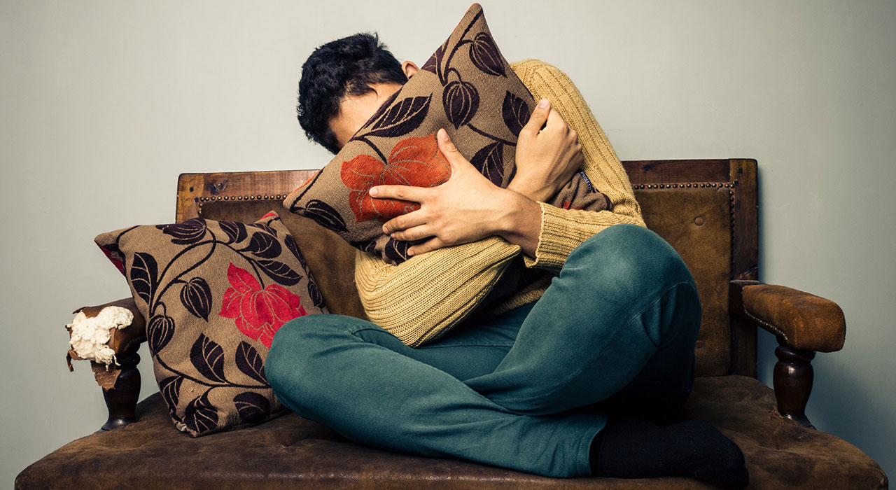 Young man is scared and hiding his face behind a cushion