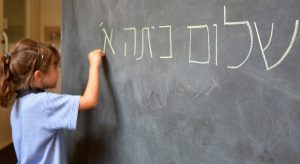 Little girl (Age 5-6) writes Hello First Grade greetings in Hebrew (Shalom Kita Alef) on a chalkboard in Israeli primary school at the beginning of the school year.