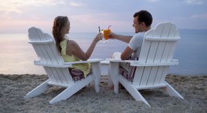 Romantic couple sitting in wooden deckchairs on the beach toasting the sunset clinking their cocktail glasses together, view from behind looking out to sea