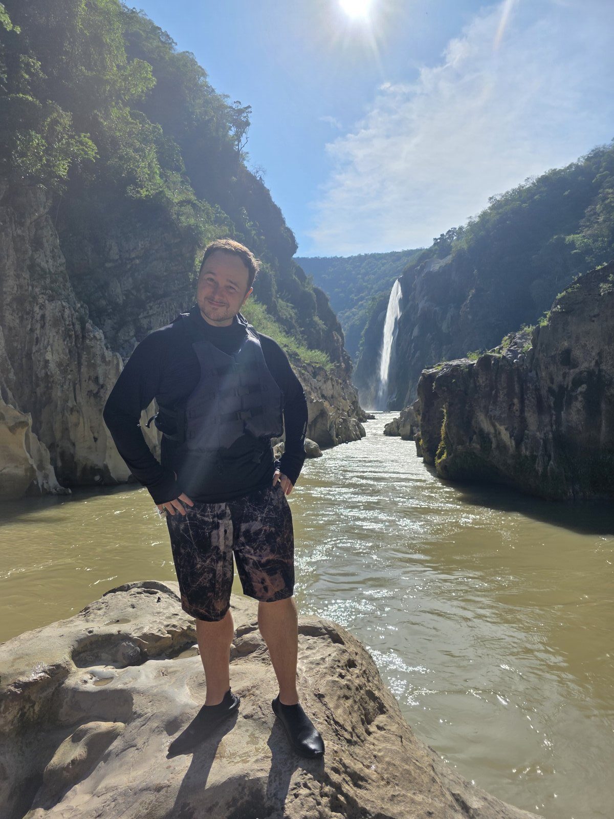 Benny standing on a rock in a canyon with the Tamul waterfall behind him in San Luis Potosi, Mexico